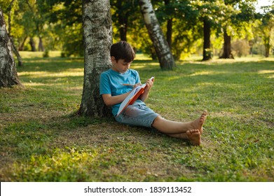 A Kid Leaning On A Tree In Park Is Holding His Journal And Writing In It .