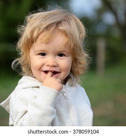 Kid little child baby boy fair-haired blond hazel-eyed smiling wearing grey hooded coat cute portrait with finger in mouth looking at camera on blurred natural background, square picture