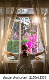 Kid Looking Through Open Window In Summer Afternoon Light. Little Girl From Behind, Opening Window To A Garden With Sun-flare.