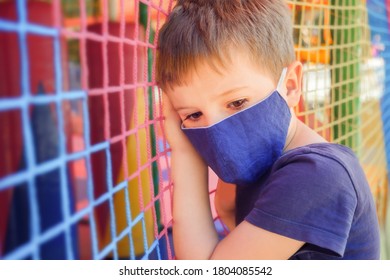 Kid In A Medical Mask Standing Near A Closed Playground. Child In Face Mask In Front Of Playground. Bored Boy In Medical Mask Near Playground During Covid-19 Global Pandemic. Coronavirus Outbreak