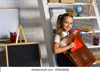 Kid Near Board And Bookshelf With Colorful Stationery On White Brick Wall Background. Pupil Puts Red Book Into Brown Leather Bag. Girl With Happy Face Gets Ready For School. Back To School Concept