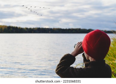Kid on lake shore watching in binoculars how flock of migrating birds training to fly in wedge