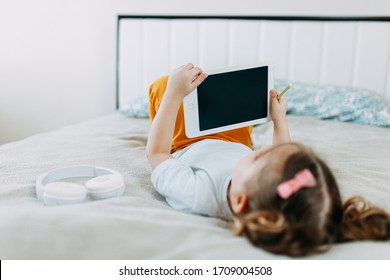 A Kid On Online Learning In The Bed, Little Girl In Bed With Tablet And Headphones, Studying From Home While Quarantine, Distance Learning