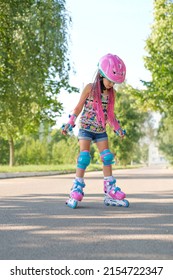 Kid On Roller Skates In Full Sports Gear Is Standing On Sidewalk Trying To Make First Move. Girl In A Pink Helmet With Afro Pigtails Learns To Skate By Doing Exercises And Practicing On Vacation