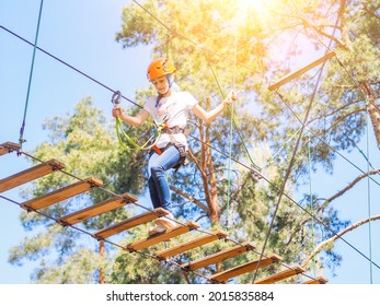 Kid In Orange Helmet Climbing In Trees On Forest Adventure Park. Girl Walk On Rope Cables And High Suspension Bridge In Adventure Summer City Park. Extreme Sport Equipment Helmet And Carabiner