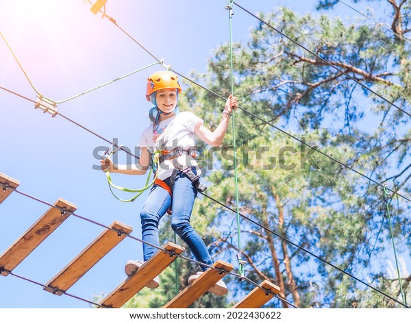 Kid in orange helmet climbing in trees on\
forest adventure park. Girl walk on rope cables and high suspension\
bridge in adventure summer city park. Extreme sport equipment\
helmet and carabiner