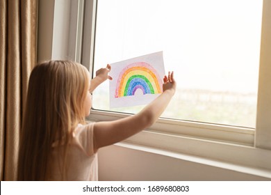 Kid painting rainbow during Covid-19 quarantine at home. Girl near window. Stay at home Social media campaign for coronavirus prevention, let's all be well, hope during coronavirus pandemic concept - Shutterstock ID 1689680803