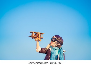 Kid Pilot With Toy Wooden Airplane Against Blue Winter Sky Background. Happy Child Playing Outdoors