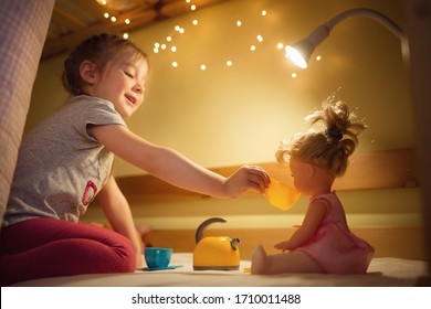 Kid Play. Girl Plays With Doll In Tea Party At Home. House With Night Lights And Lamp.