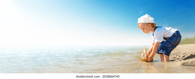 Kid play on the beach on a hot sunny day. Little girl dressed as a sailor stands barefoot on the sandy shore and launches a boat into the sea. Child dreams of travel and adventure.
