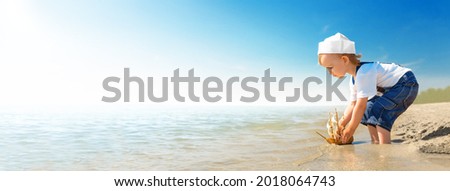 Kid play on the beach on a hot sunny day. Little girl dressed as a sailor stands barefoot on the sandy shore and launches a boat into the sea. Child dreams of travel and adventure.