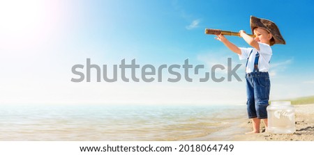 Kid play on the beach on a hot sunny day. Little girl dressed as a pirate stands barefoot on the sandy seashore with a telescope. Child with a toy fish dreams of travel and adventure.