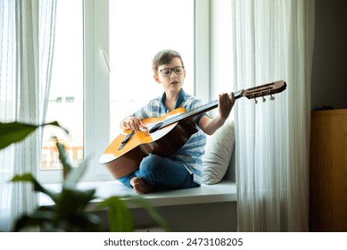 Kid playing guitar. Child boy sitting on the windowsill and playing acoustic guitar - Powered by Shutterstock