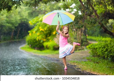 Kid playing out in the rain. Children with umbrella play outdoors in heavy rain. Little girl caught in first spring shower. Kids outdoor fun by rainy autumn weather. Child running in tropical storm.