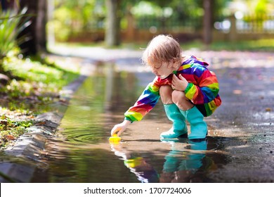 Kid Playing In The Rain In Autumn Park. Child Jumping In Muddy Puddle On Rainy Fall Day. Little Boy In Rain Boots And Red Jacket Outdoors In Heavy Shower. Kids Waterproof Footwear And Coat.