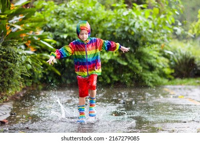 Kid Playing In The Rain In Autumn Park. Child Jumping In Muddy Puddle On Rainy Fall Day. Little Boy In Rain Boots And Rainbow Jacket Outdoors In Heavy Shower. Kids Waterproof Footwear And Coat.