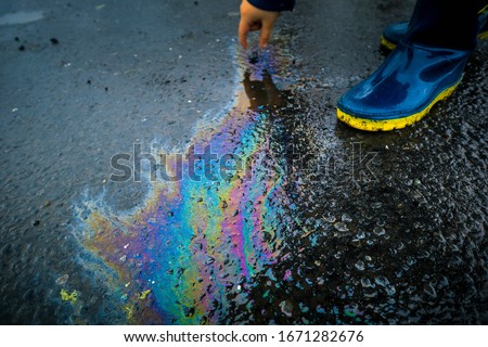  Kid playing touching the spillage and wearing boots. Petrol or petroleum spill on asphalt road. Environmental damage. Toxic oil spill in nature. Rainbow colors from chemicals.