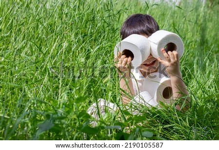 kid playing with wc toilet paper rolls tissue sits in green grass or isolated on ivory beige background.daily use product hygiene concept.child looks though paper roll hole like in binoculars.sun day