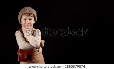 A kid plays photographer. Boy is posing with an old film camera in leather case. He is dressed in knitted retro clothes. Studio isolated on a black background. Wide image with copy space. 