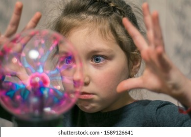 Kid Plays Witchcraft And Magic. Portrait Of A Little Girl Playing With A Plasma Ball. The Child Touches The Surface Of The Lamp Causing Lightning. Selective Focus