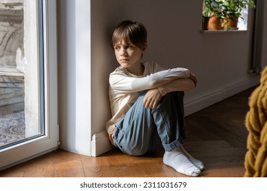 Kid psychological trauma. Lonely child boy sadly look to window hug knees sits on floor at home alone. Upset offended child thinking about family problems, bad relationship, failures with school peers - Powered by Shutterstock