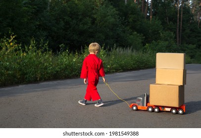 kid in a red overalls walks along the road and carries a large toy car - a truck with cardboard boxes on it. Little courier at work. Parcel delivery, little postman. be like dad. games for boys