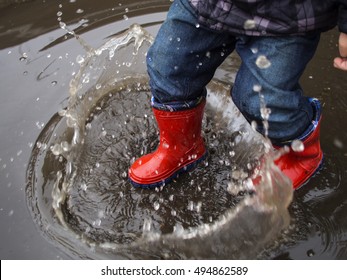 Kid In Red Rubber Boots Make Of Water Splashes In A Puddle 