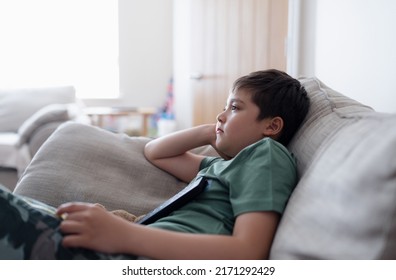 Kid With Remote Control And Looking Up With Curious Face,Young Boy Sitting On Sofa Watching Cartoon On TV, Portrait Child Lying Down On Couch Relaxing In Living Room After Back From School.