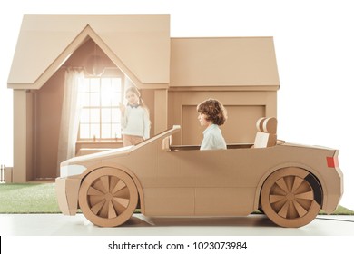 Kid Riding Cardboard Car In Front Of House While Girlfriend Waving To Him Isolated On White