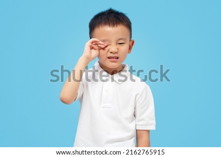 Kid rubbing eye, allergy and itching, sore eyes, wearing white shirt on blue background