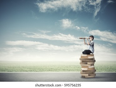 Kid of school age sitting on pile of books and looking in spyglass - Shutterstock ID 595756235