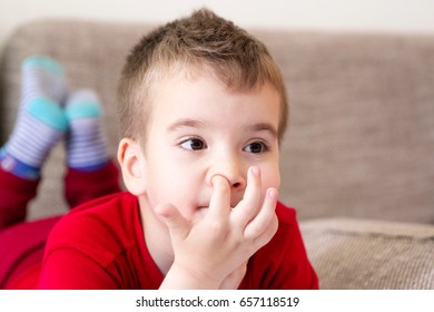 Kid Scratching A Finger In His Nose. Itching Boy Lying On His Stomach With Head In Hands And Legs Bent At The Knees While Watching TV