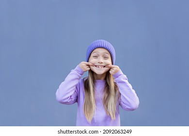 Kid Showing Missed Milk Tooth. Change Teeth Concept. Cute Little Caucasian Girl With Blonde Hair Eight Years Old On Purple Background With Copy Space. Trendy Color Of 2022 Year Very Peri