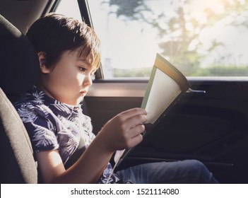 Kid Siting On Car Seat And Reading A Book, Little Boy Sitting In The Car In Child Safety Seat, Portrait Of Toddler Entertaining Himserf On A Road Trip. Concept Of Safety Taveling By Car With Children