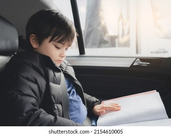 Kid Siting On Car Seat And Reading A Book, Young Boy Sitting In  Safety Seat, Portrait Of Child Entertaining Himserf On A Road Trip. Concept Of Safety Taveling By Car With Children 