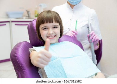 Kid Sitting In Dental Chair. Little Girl Patient Showing Thumb-up Approving Pediatric Dental Service In Modern Clinic For Children.  Focus On Happy Smiling Positively Child. Good Oral Dent Health