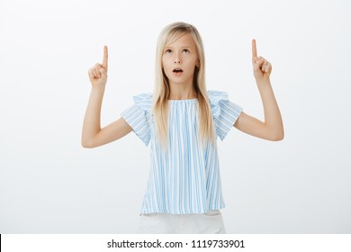 Kid Standing Still Amazed With Beehive. Shocked And Wondered Adorable Female Child In Blue Blouse, Raising Hands, Pointing And Looking Up With Interested And Curious Expression Over Gray Wall