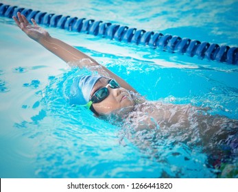 Kid swimming position backstroke. He put his hand up over the water. He wearing blue cap and goggle color black with a green strip. 