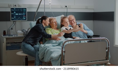 Kid taking selfie on smartphone with ill patient and family at visit in hospital ward bed. Child using mobile phone for pictures and visiting aged woman with old man and mother at clinic.