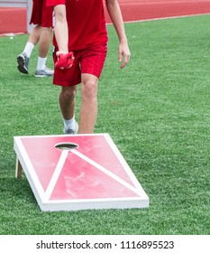 A Kid Is Throwing A Bean Bag During A Game Of Corn Hole On A Turf Field Outside.