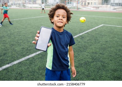 Kid Using Phone. Smiling And Excited Boy Showing Smartphone Empty Screen. Looking To Camera. Cellphone Display Mockup Mobile App Advertisement. African American Kid In Football Field. Blank Screen