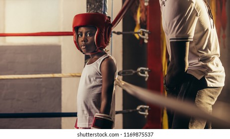 Kid Wearing Boxing Gloves And Headgear Standing In A Corner Of A Boxing Ring. Small Boy Standing Inside A Boxing Ring With His Coach Behind Him.