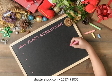 Kid Writing New Year Resolution On Black Board With Decorations And Gift Boxes, New Year And Merry Christmas Goal  Conceptual