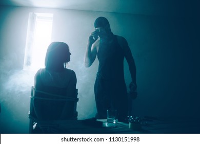 Kidnapper and his victim are in dark room with small window. He is standin in front of it and talking on the phone. Guy wears mask. She is tied to chair. Girl is looking at her kidnapper.