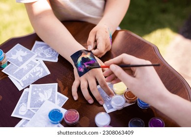 Kid's animator is creating a Shimmering sparkling glitter tattoo on a child's hand at a birthday party. Body art - Shutterstock ID 2308063409