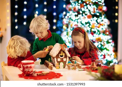 Kids Baking Christmas Gingerbread House. Children Celebrating Winter Holiday At Home. Decorated Living Room With Fireplace And Tree. Family Activity. Little Girl And Boy Making Cookies.