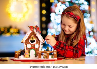 Kids Baking Christmas Gingerbread House. Children Celebrating Winter Holiday At Home. Decorated Living Room With Fireplace And Tree. Family Activity. Little Girl Making Cookies.