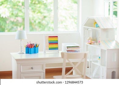 Kids Bedroom With Wooden Desk And Doll House. White Sunny Room With Big Window For Young Child. Home Interior For Little Girl. Table For Homework And Study With Lamp And Abacus, Books For School Kid.