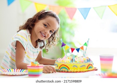 Kids Birthday Party. Children Celebrate With Colorful Cake And Gifts. Little Curly Girl Blowing Candles And Opening Birthday Presents. Friends Play With Rainbow Confetti. Party Home Decoration.