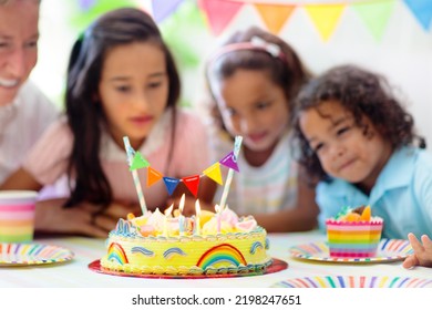 Kids Birthday Party. Children Celebrate With Colorful Cake And Gifts. Little Curly Boy Blowing Candles And Opening Birthday Presents. Friends Play With Rainbow Confetti. Party Home Decoration.